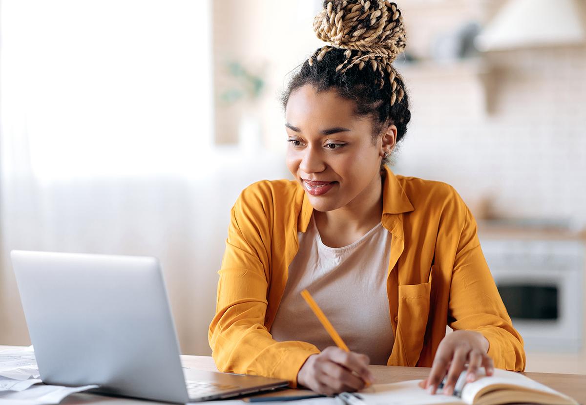 Black young woman smiling using a laptop and taking notes.