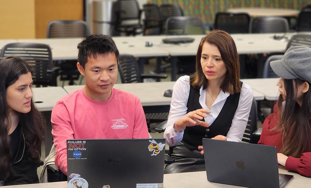 Four people seated at a classroom table collaborate using laptops while one gestures during discussion.
