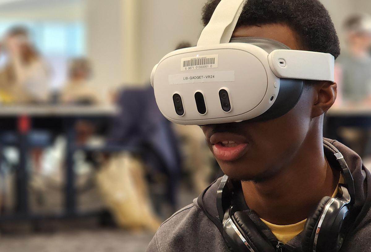 A Black young man wearing a VR headset with headphones around neck in a blurred indoor setting.